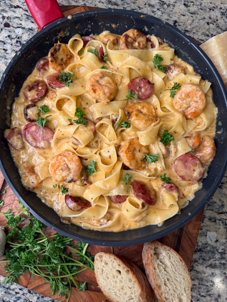 A skillet filled with creamy fettuccine pasta, shrimp, and sliced sausage, garnished with fresh parsley, served on a wooden cutting board with slices of bread beside it.