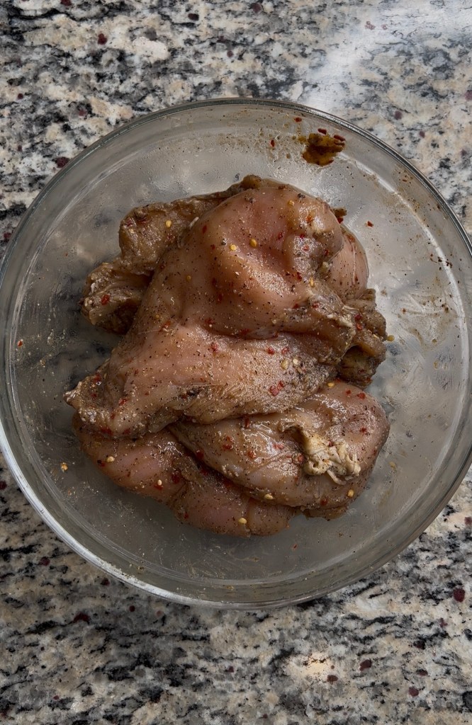 A glass bowl containing marinated chicken thighs seasoned with spices, placed on a granite countertop.