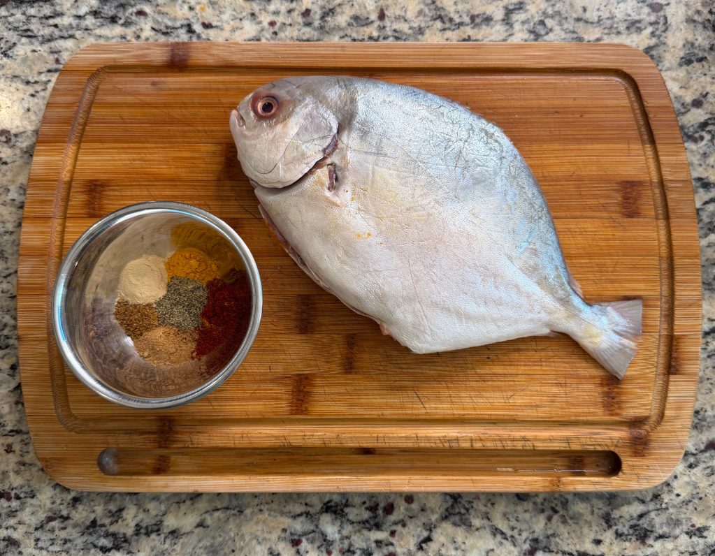 A whole fish laid on a wooden cutting board next to a bowl of various spices, showcasing ingredients for a meal preparation.