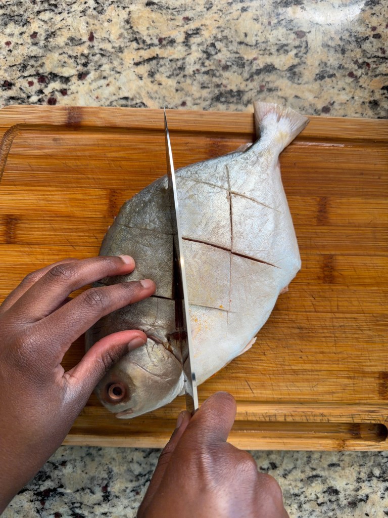 A person slicing through a whole fish on a wooden cutting board, with a marble countertop in the background.