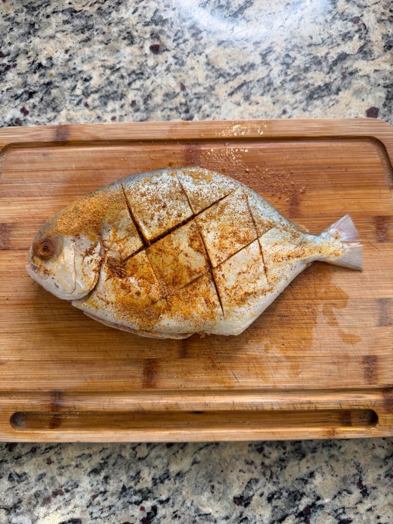 A whole fish seasoned with spices, diagonally scored, resting on a wooden cutting board.