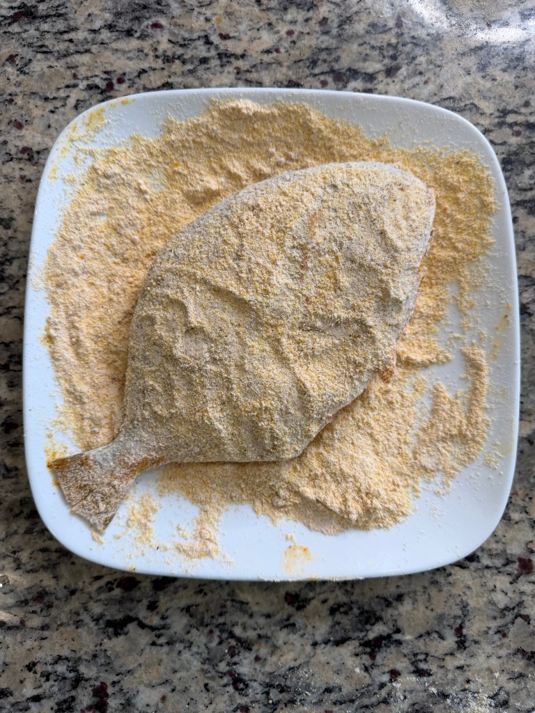 A white plate covered with cornmeal and a pompano coated in a dry batter, resting on a granite countertop.