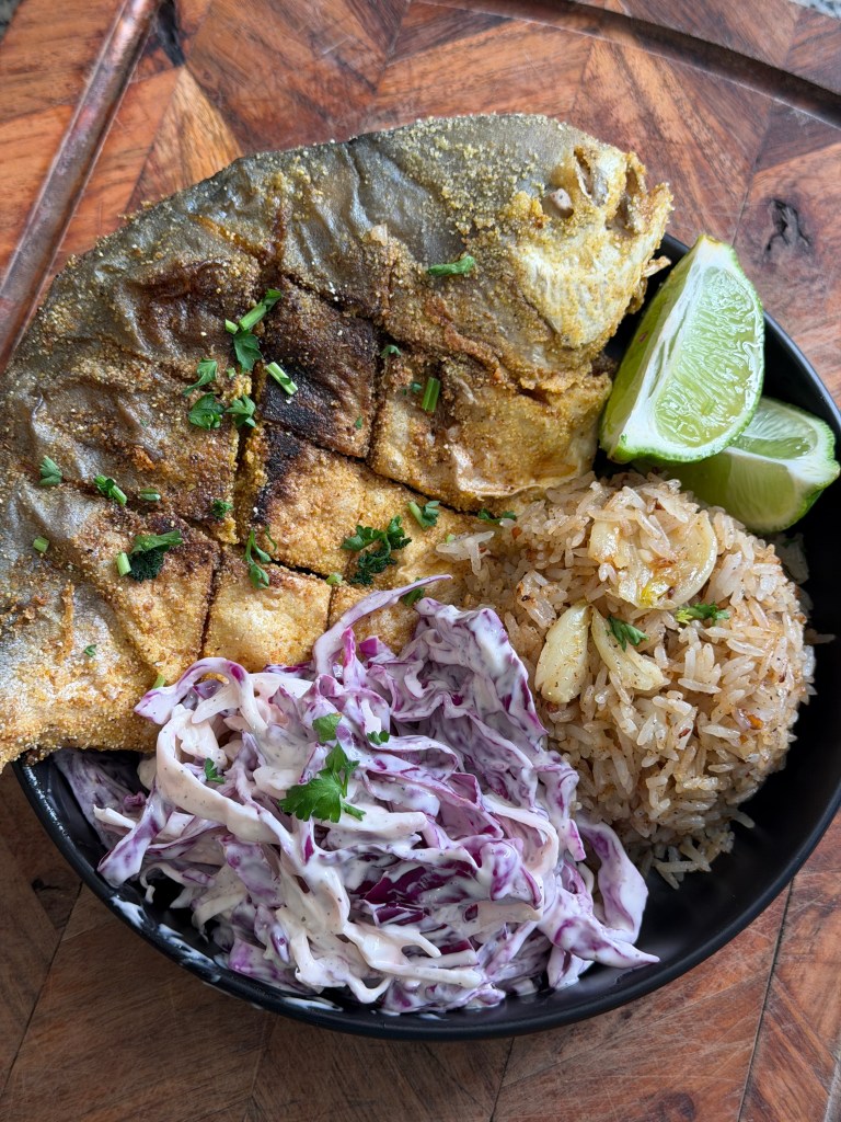A plate featuring a golden-brown grilled fish garnished with herbs, accompanied by fluffy rice with garlic, and a side of creamy purple and white coleslaw, and fresh lime wedges.