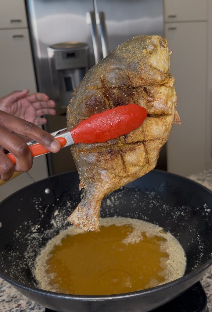 A hand holding a spatula lifts a fried pompano from a frying pan filled with oil.