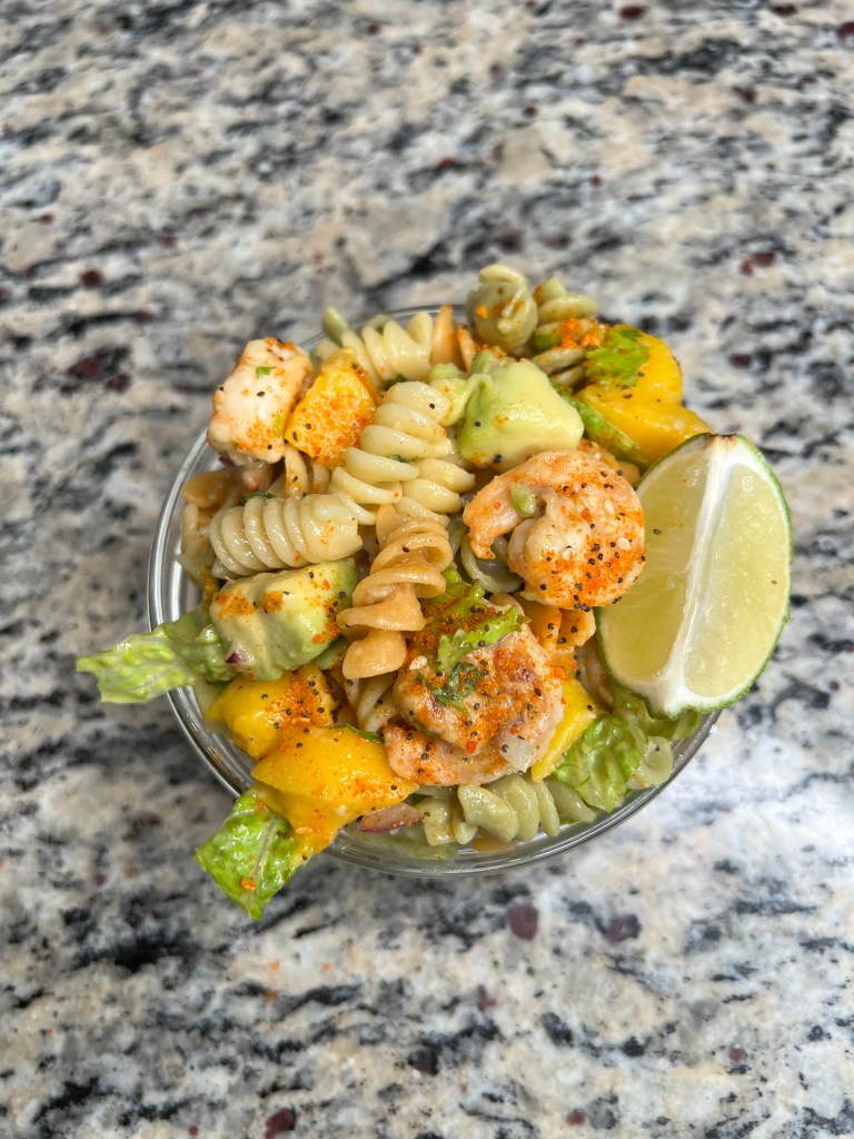 A bowl of shrimp pasta salad featuring rotini pasta, mango chunks, avocado, and lettuce, garnished with lime wedges.