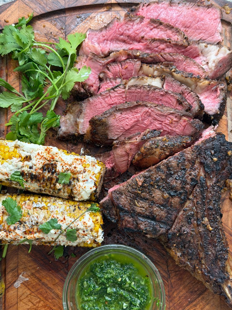 A close-up of sliced beef steaks with a green herb garnish, accompanied by grilled corn topped with cheese and spices, and a small bowl of green sauce on a wooden cutting board.