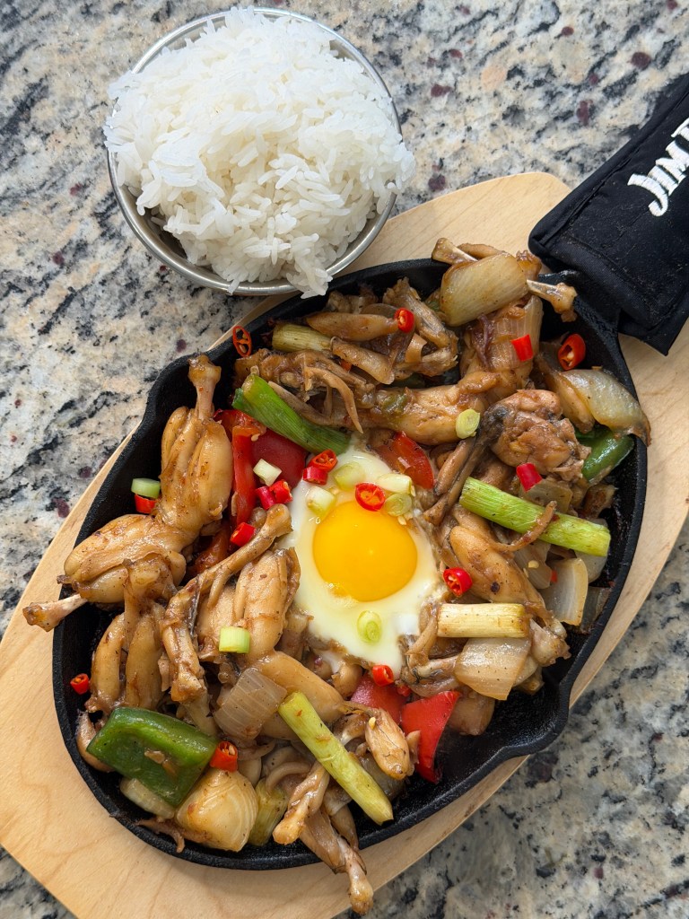 A sizzling hot plate of stir-fried chicken feet with vegetables, topped with a fried egg and sliced chili peppers, served alongside a bowl of steamed white rice.