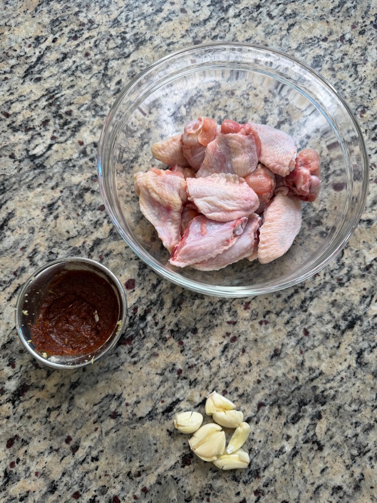 A glass bowl containing raw chicken wings placed on a granite countertop, alongside a small bowl of red marinade and several cloves of garlic.