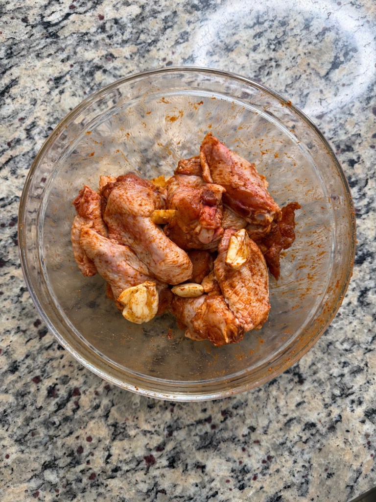 A glass bowl containing marinated chicken wings with spices and garlic, placed on a granite countertop.