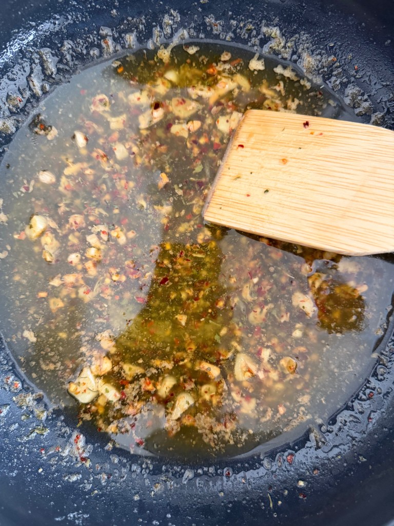 A close-up of a frying pan with a mixture of chopped garlic, herbs, and spices simmering in oil, along with a wooden spatula stirring the ingredients.