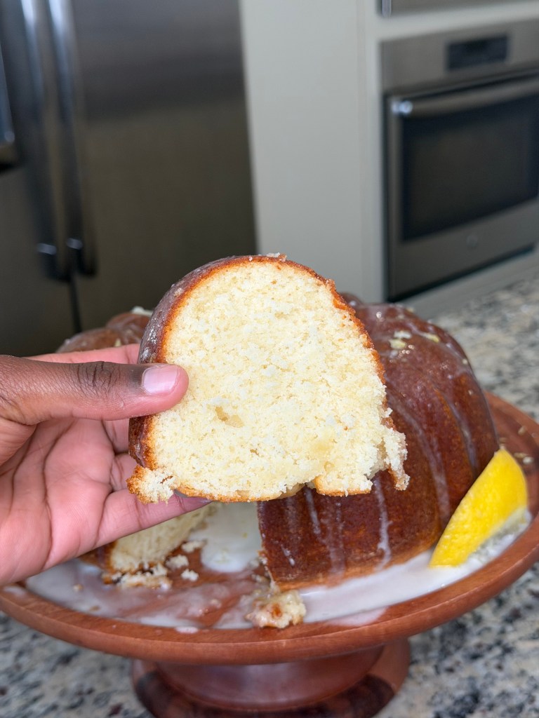 A hand holding a slice of moist lemon bundt cake with a bundt cake on a plate in the background, with a slice cut out and some glaze drizzling down.