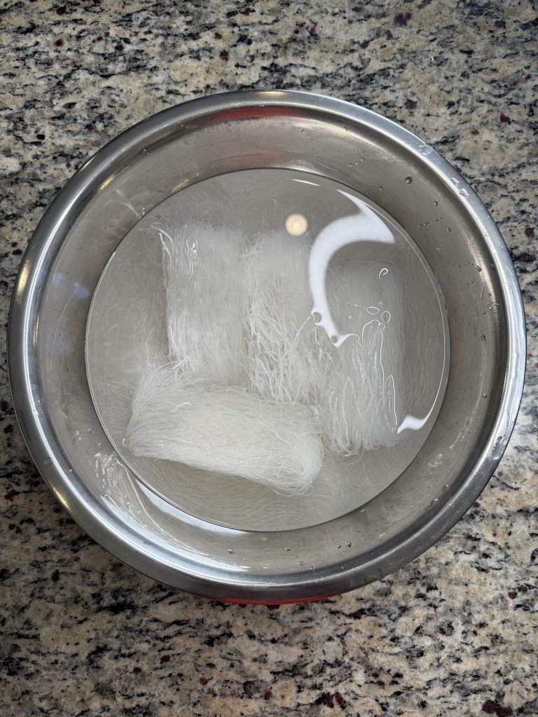 A bowl of water containing soaked transparent rice noodle sheets, placed on a granite countertop.