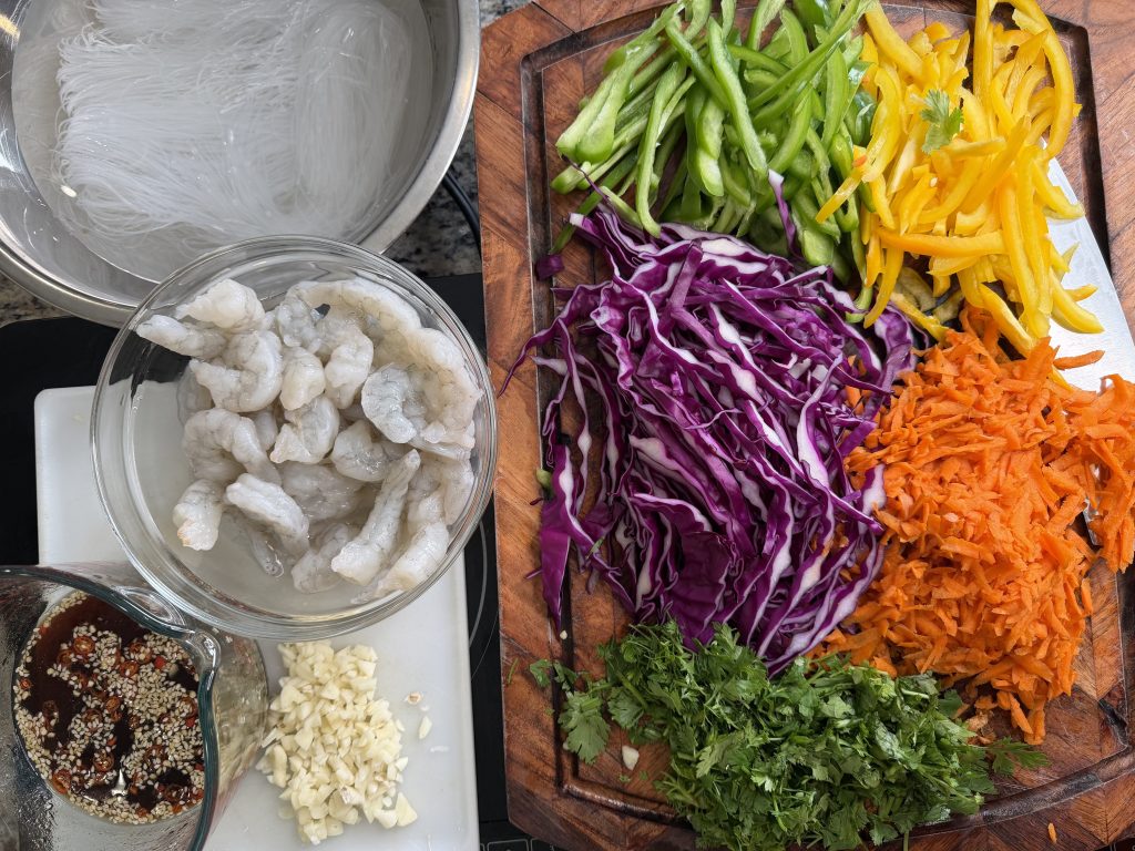 A bowl of raw shrimp alongside a bowl of soaked rice noodles, with a variety of colorful vegetables including shredded purple cabbage, sliced green and yellow bell peppers, and grated carrots on a wooden cutting board. There is also a small bowl of sauce with sesame seeds and minced garlic at the bottom.