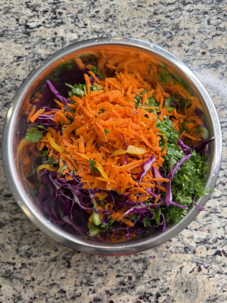 Bowl of mixed shredded vegetables including orange carrots, purple cabbage, and green herbs on a marble countertop.