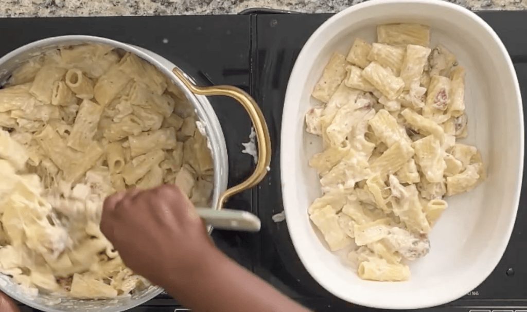 A close-up of a person stirring a large pot of creamy pasta, with rigatoni pasta mixed in a sauce, alongside a baking dish filled with similar pasta.