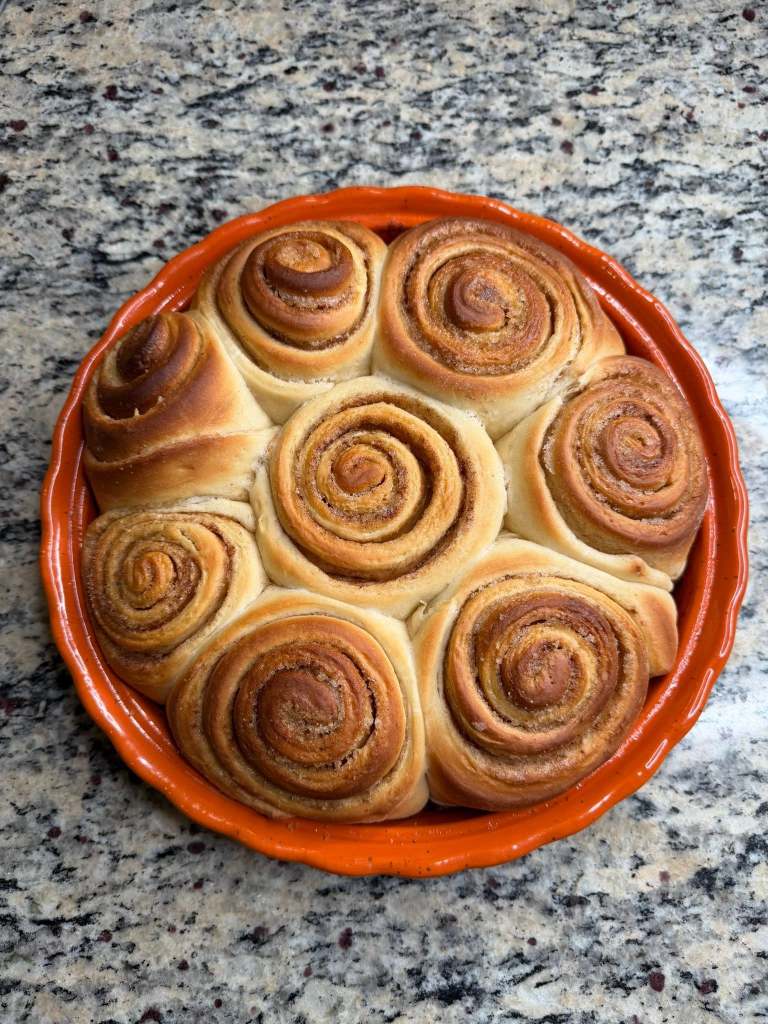 A freshly baked cinnamon roll cake arranged in a circular pattern in an orange ceramic dish, on a granite countertop.