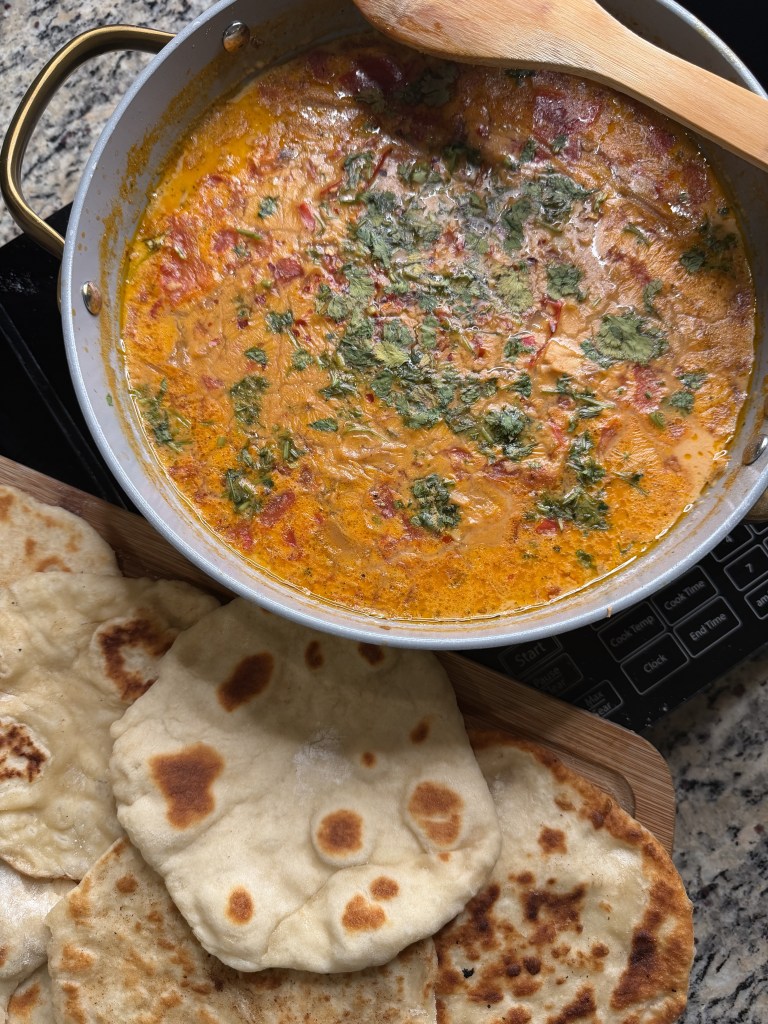 A delicious, creamy curry dish with herbs and spices in a pan, accompanied by freshly baked naan bread arranged on a wooden board.