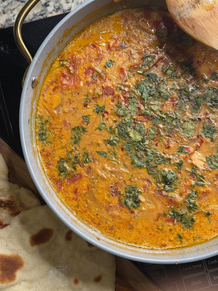 A close-up of a creamy sauce with diced tomatoes and fresh herbs, simmering in a gray pot. Soft, flatbread is visible on a wooden surface beside the pot.