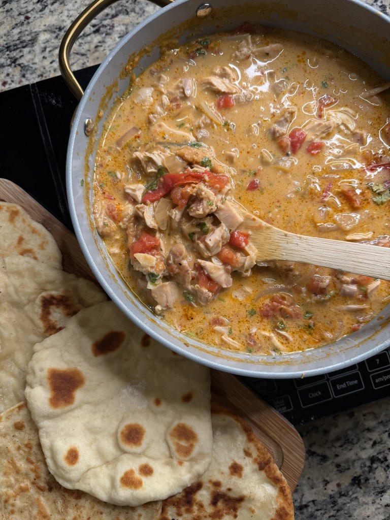 A pot filled with a creamy chicken and tomato stew, garnished with herbs, alongside freshly baked flatbreads on a wooden surface.