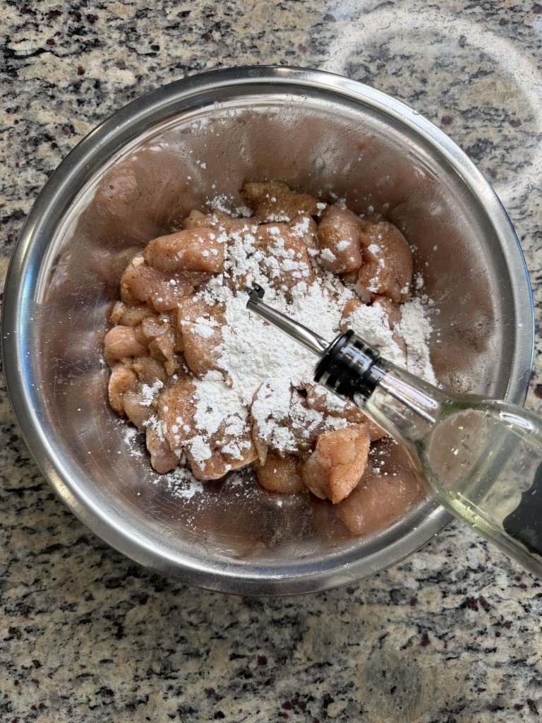 Raw chicken pieces in a stainless steel bowl being seasoned with flour and oil.