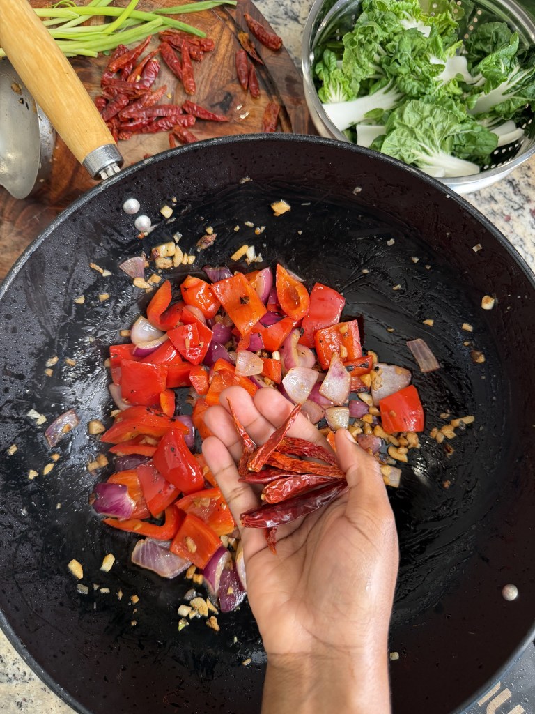 A hand holding dried red chili peppers above a wok filled with diced red bell peppers and onions, sautéing with garlic in oil.