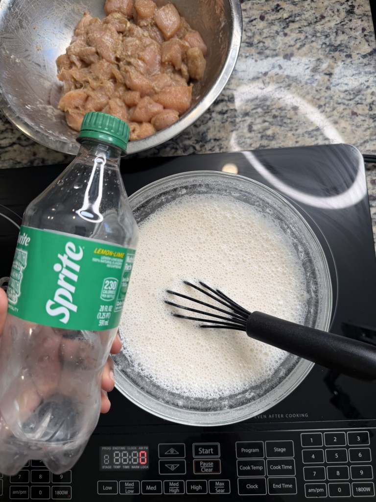 A hand holding a bottle of Sprite above a bowl of foamy ingredients, with a bowl of diced marinated chicken in the background. The scene is set on a kitchen countertop with an electric stove displaying the timer.