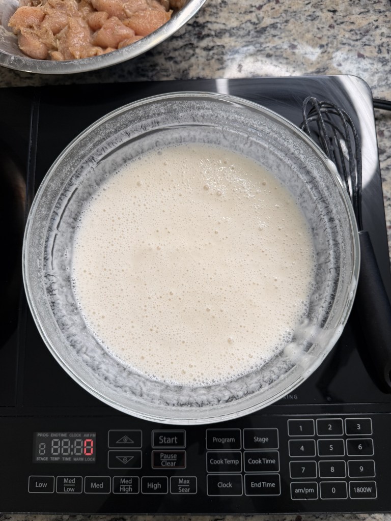 A glass bowl filled with a frothy liquid mixture, placed on a digital stovetop. In the background, a stainless steel bowl contains diced chicken.
