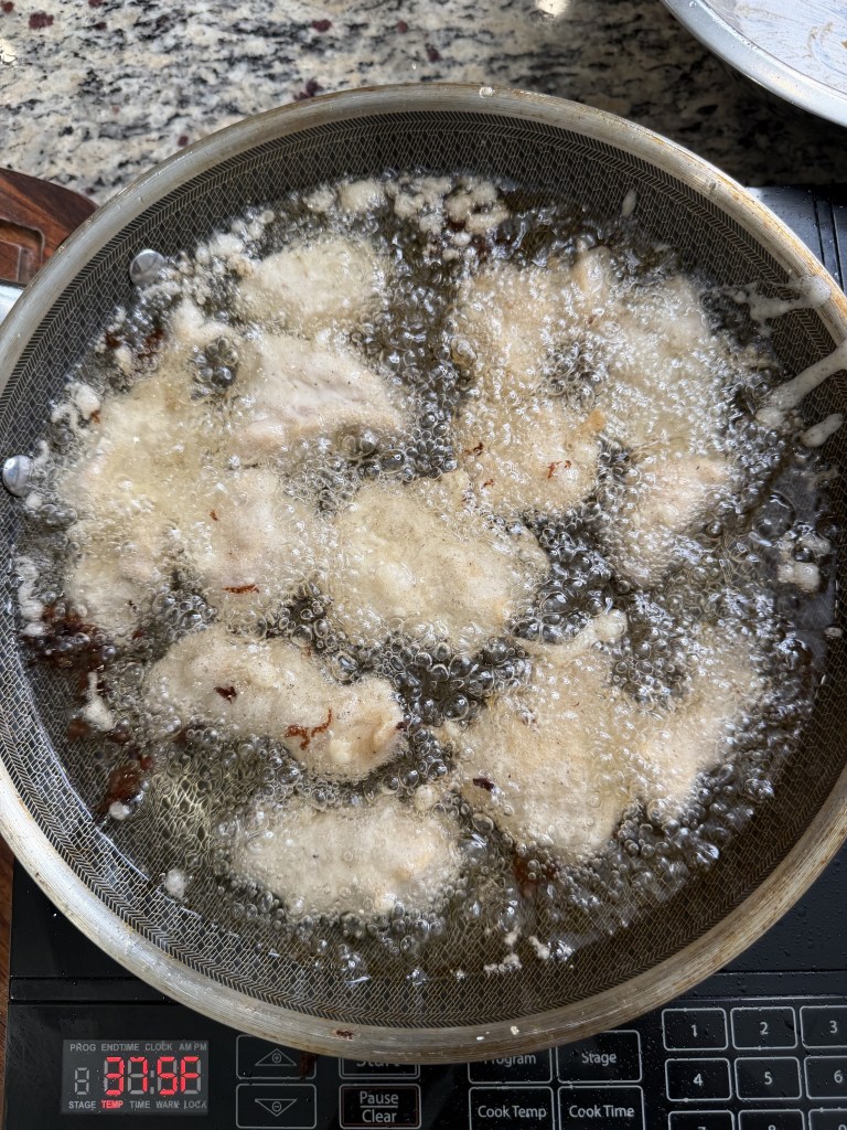 Close-up of sizzling batter-fried chicken in hot oil, with bubbles rising in a strainer over a digital cooking timer.
