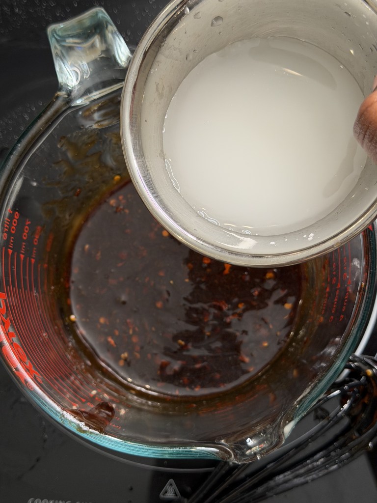 A measuring cup containing dark sauce with spices beside a small bowl of clear liquid, placed on a black countertop.