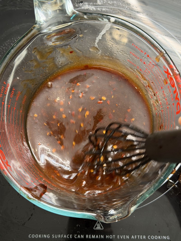 A clear measuring cup containing a dark, glossy sauce with red pepper flakes, being whisked with a metal whisk on a cooking surface.