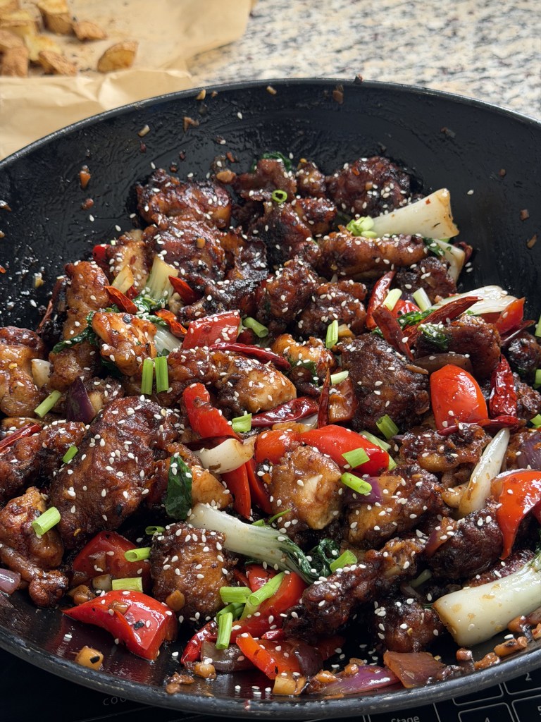 A close-up image of a wok filled with stir-fried chicken and colorful vegetables, including red bell peppers, green onions, bok choy, and sesame seeds, on a textured surface.