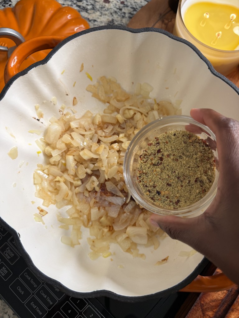 A hand pouring seasoning into a pot with sautéed onions on a stovetop, with an orange pumpkin decoration and a candle in the background.