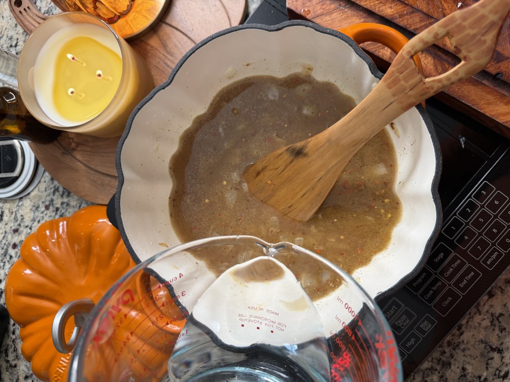 A pot with a brown sauce or broth in it, stirred with a wooden spoon, surrounded by a measuring cup with water, a lit candle, and a decorative orange pumpkin-shaped dish on a kitchen counter.