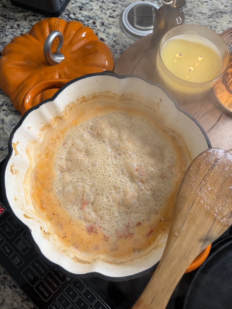 A bubbling mixture in a white frying pan on a countertop, surrounded by kitchen items including an orange pumpkin-shaped dish, a glass candle, and a wooden spoon.