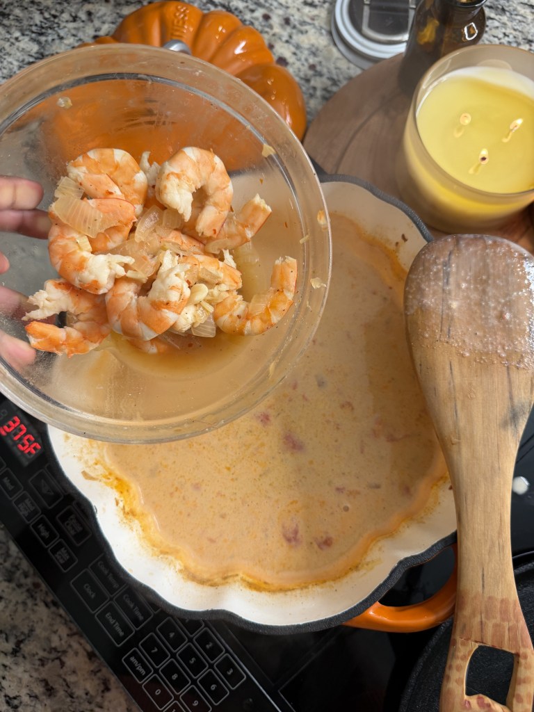 A person holding a bowl of cooked shrimp above a simmering pot of seafood soup on a kitchen countertop.