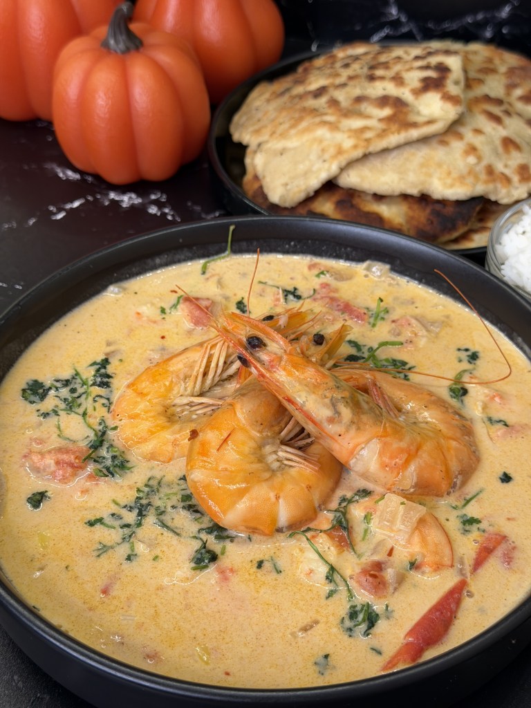 A bowl of creamy shrimp curry with large shrimp, chunks of tomatoes, and green herbs, served alongside flatbreads and decorative pumpkins.