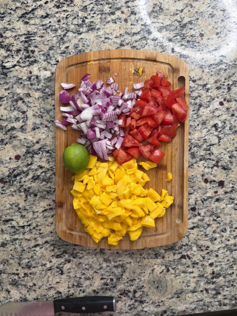Chopped red onions, diced tomatoes, and diced mango on a wooden cutting board with a lime, set on a granite countertop.