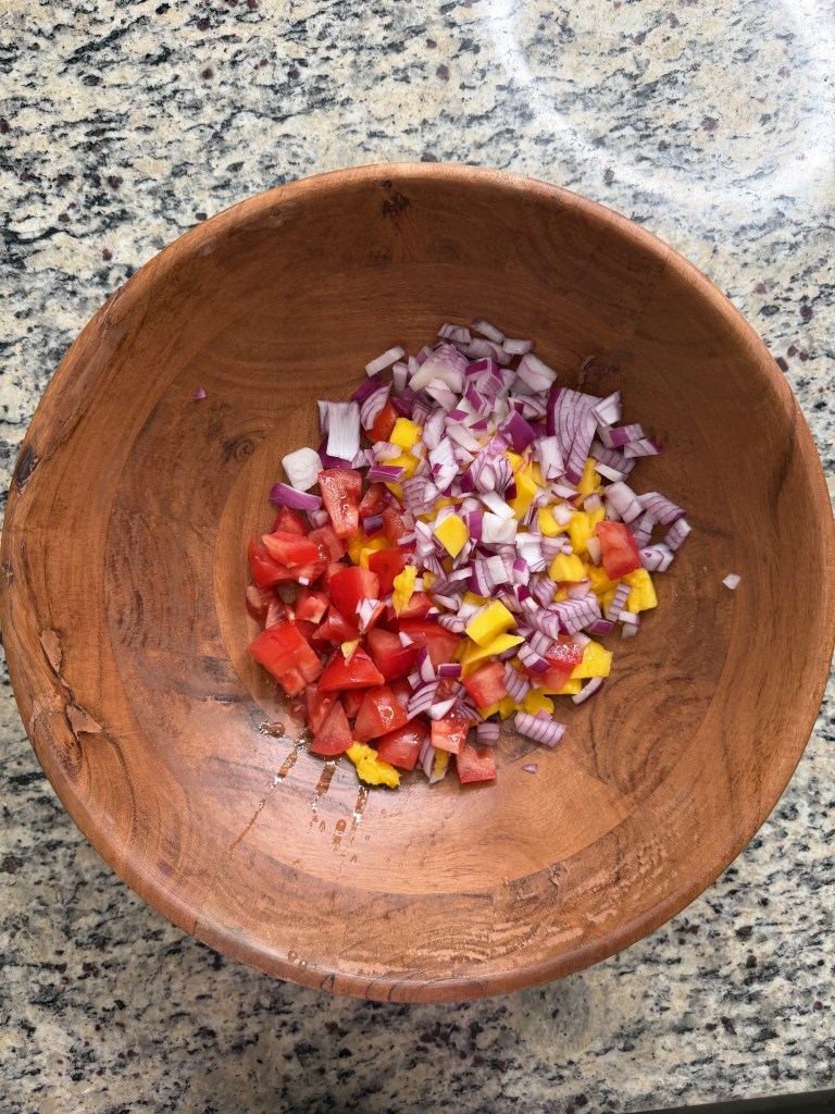 Chopped tomatoes, red onions, and yellow bell peppers in a wooden bowl on a granite countertop.