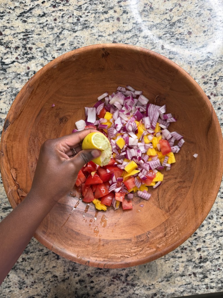 A person squeezing a lemon over a wooden bowl filled with chopped tomatoes, red onions, and yellow bell peppers on a granite countertop.