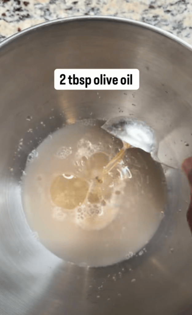 A metal mixing bowl containing a mixture with 2 tablespoons of olive oil being stirred.