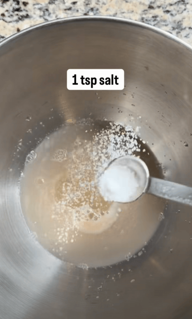 A metal mixing bowl containing water with a teaspoon of salt being added.