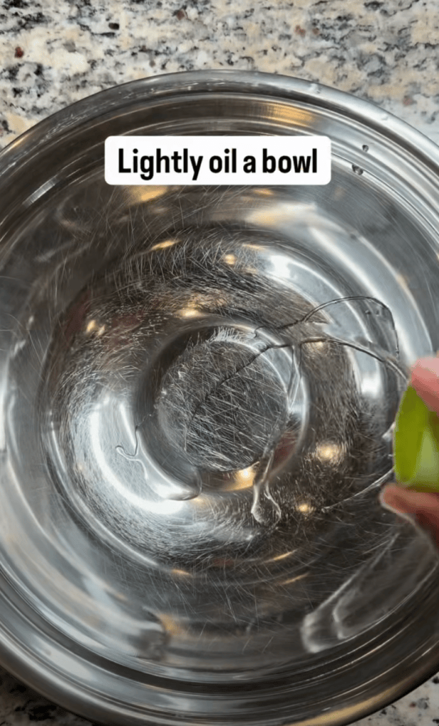 A hand gently oils the interior of a metallic mixing bowl, with a text overlay reading 'Lightly oil a bowl'.