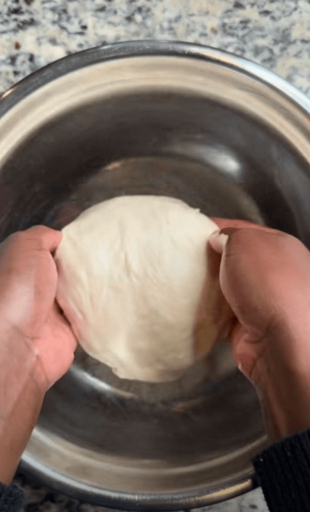 Hands kneading dough in a stainless steel bowl on a marble countertop.