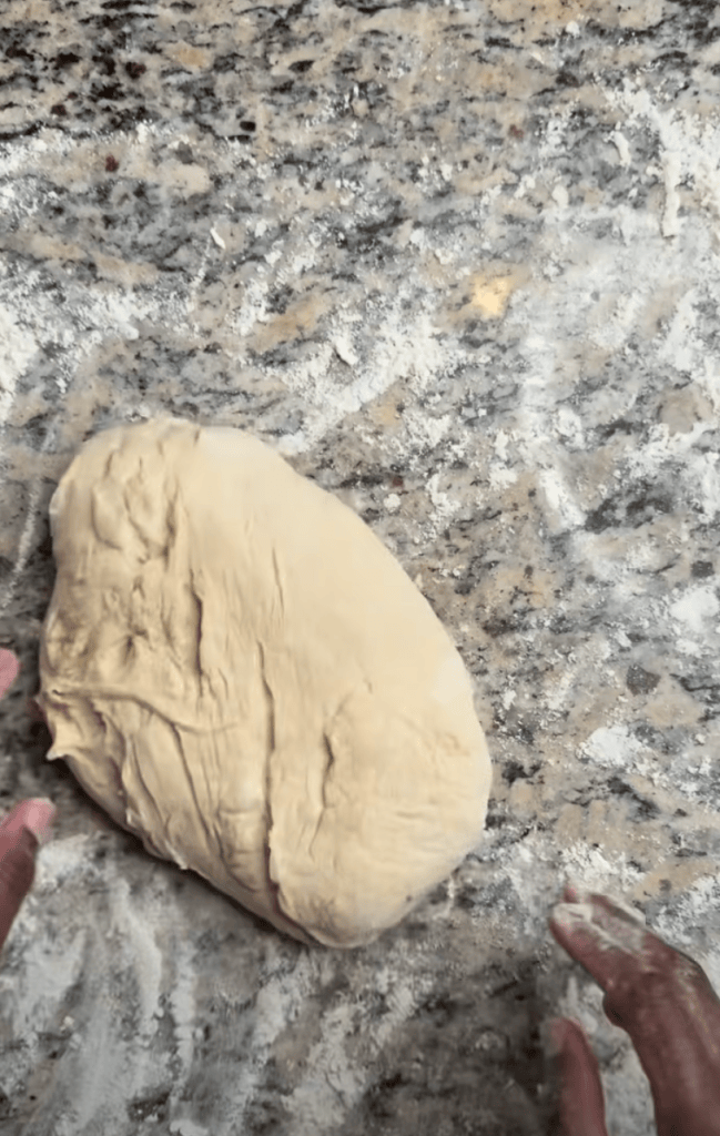 A close-up view of dough resting on a floured granite countertop, with hands positioned nearby.
