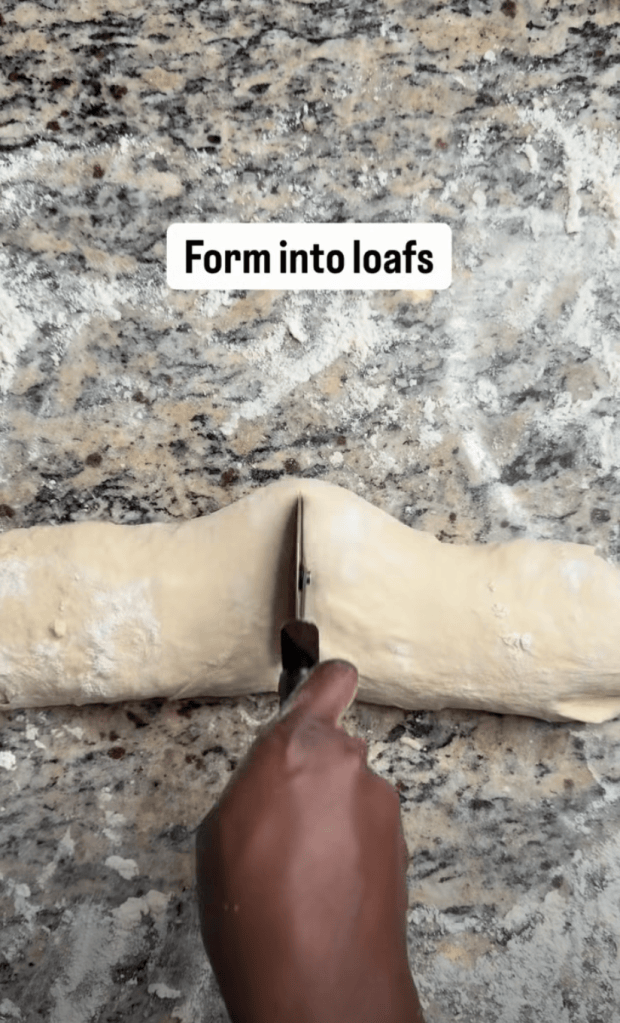 A hand holding a knife cutting dough into two loaves on a floured countertop.