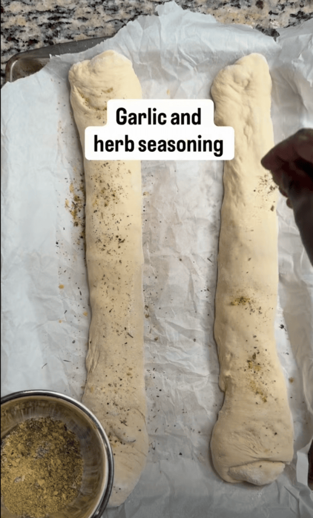 Two pieces of dough on parchment paper, with garlic and herb seasoning sprinkled on top, and a bowl of seasoning in the foreground.