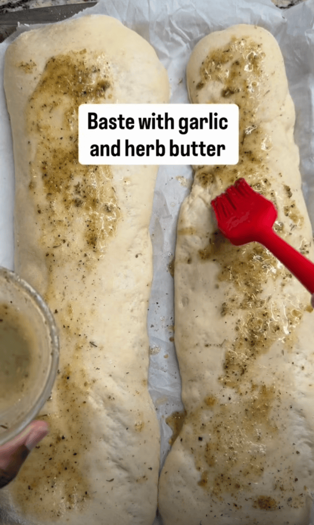 Two pieces of dough being basted with garlic and herb butter using a red brush, with a bowl of butter visible in the hand of the person.