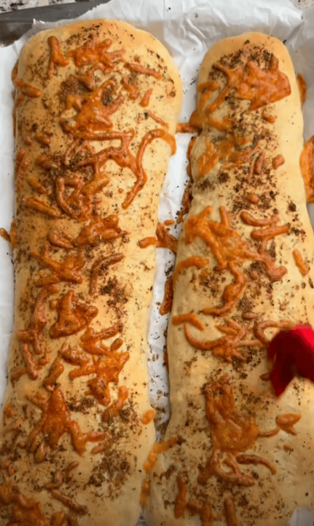 Two freshly baked loaves of bread topped with melted cheese and seasoning on parchment paper.