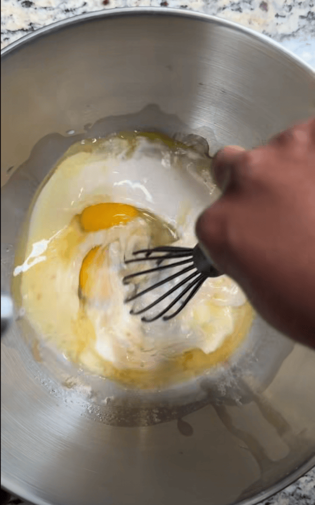 A hand whisking eggs and flour in a stainless steel bowl on a marble countertop.