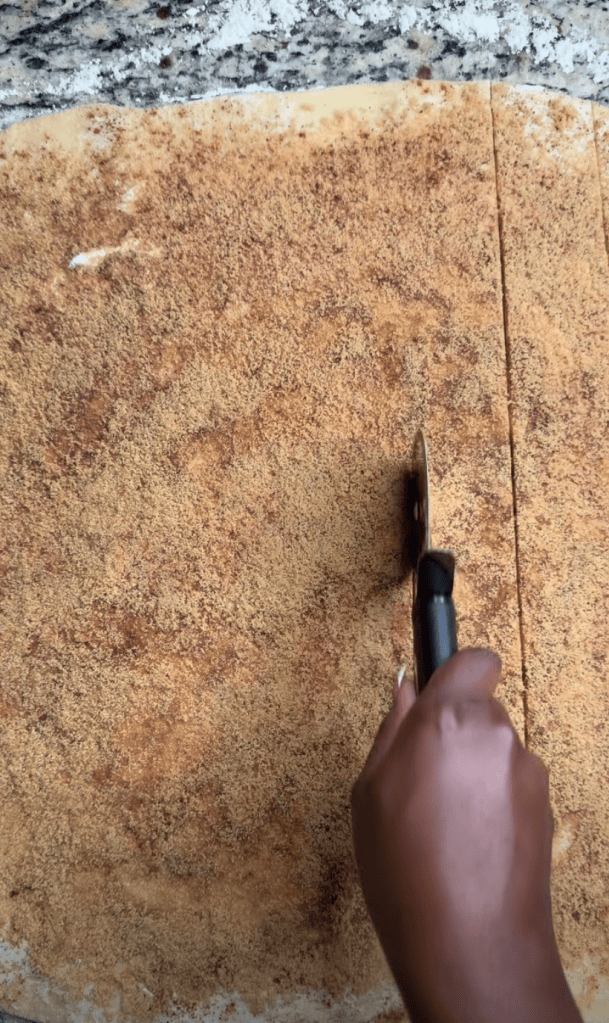 A hand using a rolling cutter to slice through a cinnamon-sugar topped dough on a countertop.